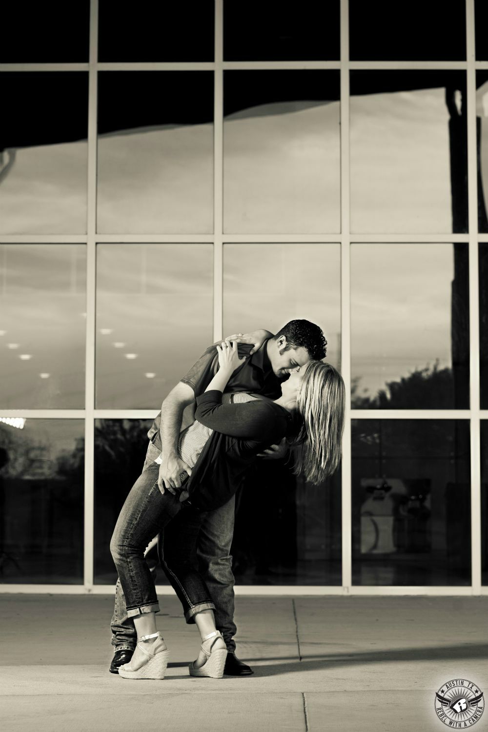 Buxom blond girl wearing an unbuttoned  dark sweater over a light blouse and short blue jeans and tan wedges is dipped by a dark haired guy in a dark grey short sleeve button up shirt and light colored jeans in front of a glass wall at the Palmer Events Center in thie fanciful engagement photo in Austin.  